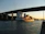 A large cargo ship carrying numerous containers sails under a tall bridge spanning a calm waterway. The lighting suggests late afternoon or early evening, with the sky showing tones of orange and blue. On the horizon, there are industrial buildings and trees along the riverbank.
