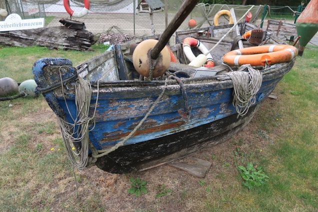 A weathered wooden boat with peeling blue and black paint rests on grass, surrounded by nautical equipment like lifebuoys, ropes, and fishing gear. Signs of aging and wear are evident, giving it a rustic appearance. Behind the boat, a fence and various maritime items add to the setting.