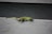 A small, colorful gecko with a vibrant green and subtle red pattern stands on a textured gray wooden surface. The lizard has a long tail and its eyes are prominently visible, suggesting attentiveness as it basks quietly.