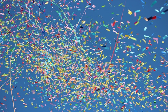A vibrant explosion of colorful confetti against a clear blue sky. The confetti consists of various shapes and sizes, creating a dynamic and festive pattern. Bright reds, yellows, greens, and blues dominate the scene.