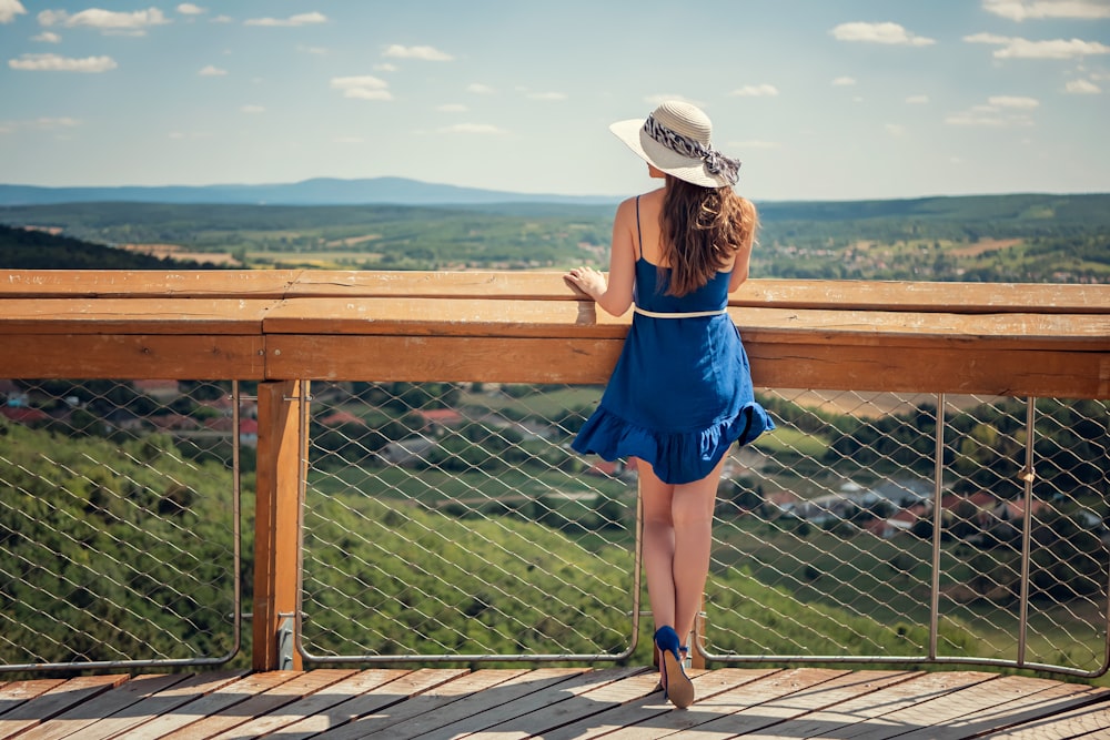 Mujer con vestido azul sin mangas con sombrero blanco de pie en el puente de madera marrón durante el día