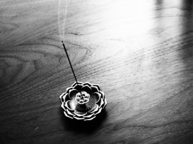 A detailed incense holder resembling a lotus flower is placed on a wooden surface. The holder has a patterned design and holds a burning incense stick, with delicate smoke trails rising upwards. The lighting accentuates the grain of the wood, creating a serene and minimalistic composition.