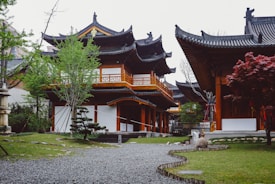 Traditional East Asian-style architecture features prominently with intricate wooden structures, ornate roofs, and detailed carvings. The scene includes lush greenery with manicured trees, shrubs, and a pathway made of stones leading through a well-maintained garden.