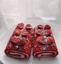 Two slices of a red velvet roll cake are placed on a white plate. The cake is topped with slices of fresh strawberries, white chocolate balls, and sprinkled with shredded coconut. The background is blurred, creating a soft focus effect.