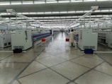 A spacious, well-lit industrial setting with numerous large white machines lined up in parallel rows. A person in uniform appears to be operating one of the machines. Red and blue bins are placed at intervals along the line of machines, which appear to be automated textile machines. The facility is clean and modern, with the floor marked with geometric patterns.