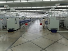 A spacious, well-lit industrial setting with numerous large white machines lined up in parallel rows. A person in uniform appears to be operating one of the machines. Red and blue bins are placed at intervals along the line of machines, which appear to be automated textile machines. The facility is clean and modern, with the floor marked with geometric patterns.