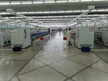 A spacious, well-lit industrial setting with numerous large white machines lined up in parallel rows. A person in uniform appears to be operating one of the machines. Red and blue bins are placed at intervals along the line of machines, which appear to be automated textile machines. The facility is clean and modern, with the floor marked with geometric patterns.