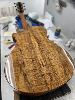 A beautifully crafted guitar body with a polished wooden surface featuring rich, intricate grain patterns. The guitar is placed on a workbench surrounded by various tools and materials.