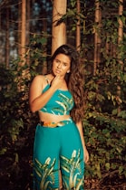 A woman with long hair stands confidently in a forest, wearing a blue-green outfit with nature-inspired patterns. The setting sun casts a warm glow, highlighting her features and the surrounding foliage.