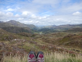 A pair of hiking shoes is visible at the bottom of the image, resting on a grassy hillside with a vast, mountainous landscape stretching into the distance. The sky is mostly cloudy with patches of blue peeking through, and the terrain is a mix of green and brown tones, suggesting late summer or early autumn. The mountains in the background are rolling and expansive, covered in vegetation.