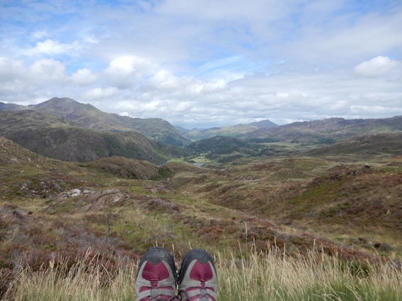 A pair of hiking shoes is visible at the bottom of the image, resting on a grassy hillside with a vast, mountainous landscape stretching into the distance. The sky is mostly cloudy with patches of blue peeking through, and the terrain is a mix of green and brown tones, suggesting late summer or early autumn. The mountains in the background are rolling and expansive, covered in vegetation.