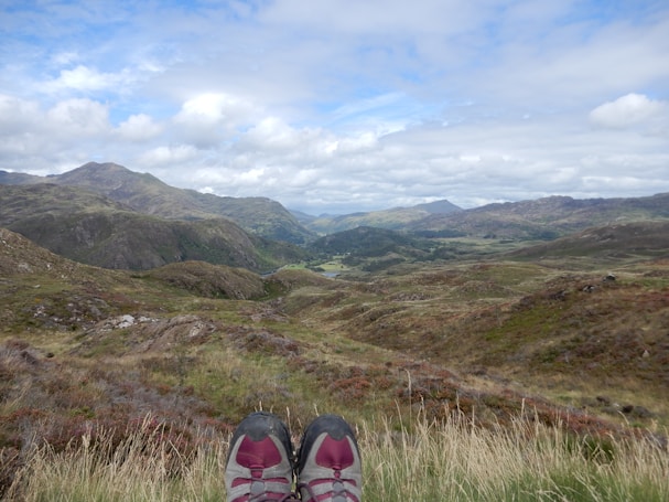 A pair of hiking shoes is visible at the bottom of the image, resting on a grassy hillside with a vast, mountainous landscape stretching into the distance. The sky is mostly cloudy with patches of blue peeking through, and the terrain is a mix of green and brown tones, suggesting late summer or early autumn. The mountains in the background are rolling and expansive, covered in vegetation.