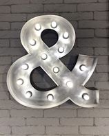 A large, metallic ampersand symbol with embedded round lights is mounted on a textured white brick wall. The symbol emits a bright glow, contrasting sharply with the rustic background.
