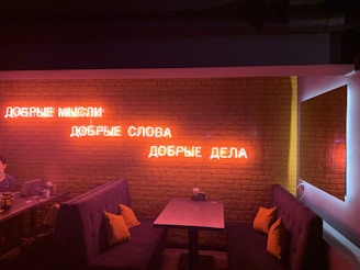 A dimly lit room with a brick wall features neon Russian text glowing in red. The interior presents two cozy booth-style seating areas with dark cushions and yellow pillows. There's a small table between the seats, and a person sits at the table on the left, with a laptop.