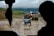 A muddy rural road is occupied by a small blue three-wheeled vehicle filled with several people wearing colorful clothes. Ahead, a truck is moving slowly, and the background consists of rolling hills and dark clouds, suggesting an overcast day. The foreground is silhouetted by parts of a vehicle interior, indicating the perspective from inside another vehicle.