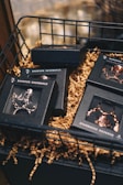 An assortment of small black boxes with transparent lids is arranged in a wire basket. The boxes contain items that appear to be chains or jewelry, and they are surrounded by crinkled brown paper filler. The setting has a warm, inviting atmosphere with soft focus in the background.