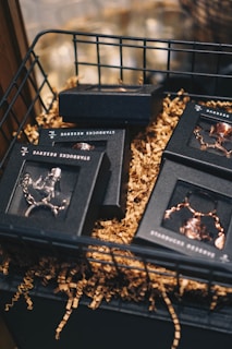 An assortment of small black boxes with transparent lids is arranged in a wire basket. The boxes contain items that appear to be chains or jewelry, and they are surrounded by crinkled brown paper filler. The setting has a warm, inviting atmosphere with soft focus in the background.