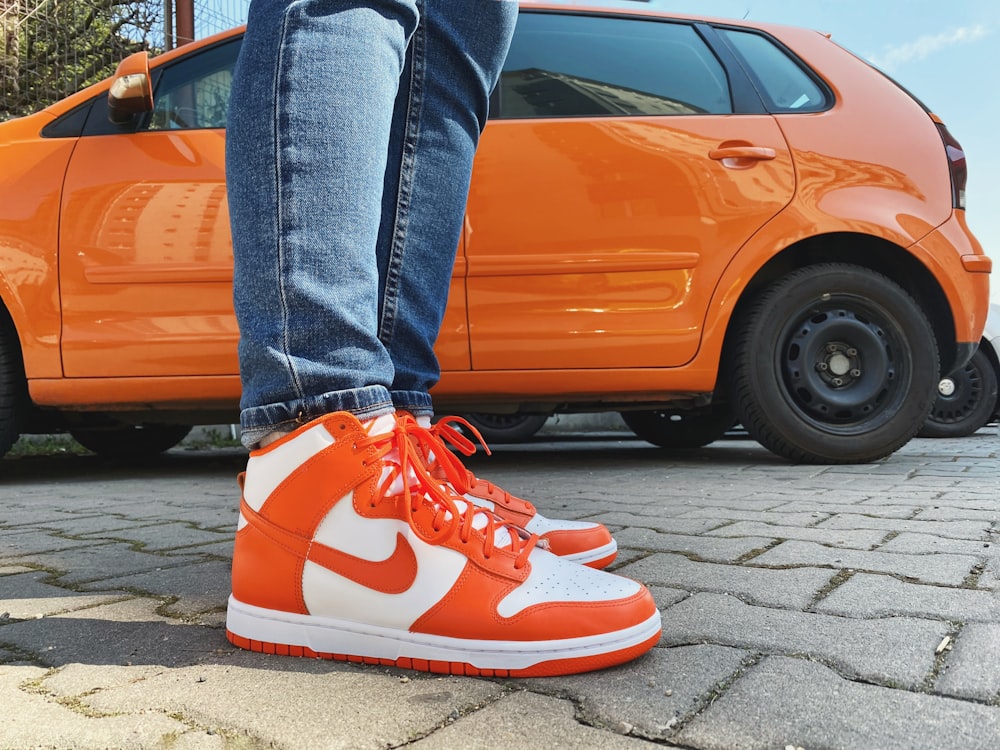 Person in blauen Jeans und roten Nike-Sneakers neben orangefarbenem Auto