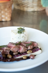 A gourmet dish consisting of sliced medium-rare beef with a side of rice topped with herbs, garnished with olives and leafy vegetables, elegantly arranged on a white plate. The background features a woven basket blurred out.