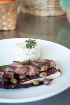 A gourmet dish consisting of sliced medium-rare beef with a side of rice topped with herbs, garnished with olives and leafy vegetables, elegantly arranged on a white plate. The background features a woven basket blurred out.
