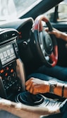 A driver's hand is gripping the gear shift of a car, with another hand on the steering wheel. The car's interior features a modern dashboard with various controls and a digital display screen. Sunlight streams through the window, highlighting the details of the car's design.