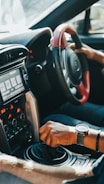 A driver's hand is gripping the gear shift of a car, with another hand on the steering wheel. The car's interior features a modern dashboard with various controls and a digital display screen. Sunlight streams through the window, highlighting the details of the car's design.
