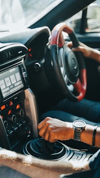 A driver's hand is gripping the gear shift of a car, with another hand on the steering wheel. The car's interior features a modern dashboard with various controls and a digital display screen. Sunlight streams through the window, highlighting the details of the car's design.