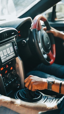 A driver's hand is gripping the gear shift of a car, with another hand on the steering wheel. The car's interior features a modern dashboard with various controls and a digital display screen. Sunlight streams through the window, highlighting the details of the car's design.