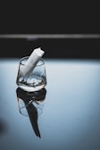 A transparent glass is placed on a reflective surface, slightly tilted with a white rolled-up paper inside. The background is dark, enhancing the contrast with the glass and paper.