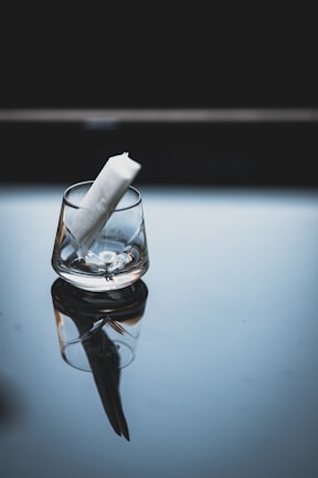 A transparent glass is placed on a reflective surface, slightly tilted with a white rolled-up paper inside. The background is dark, enhancing the contrast with the glass and paper.