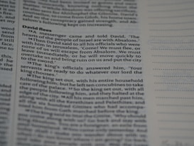 Close-up view of a book page with text in a structured format, resembling a biblical or literary passage. The text includes a narrative involving characters and dialogue. The page layout includes a header and numbered lines, suggesting a divided section of content.