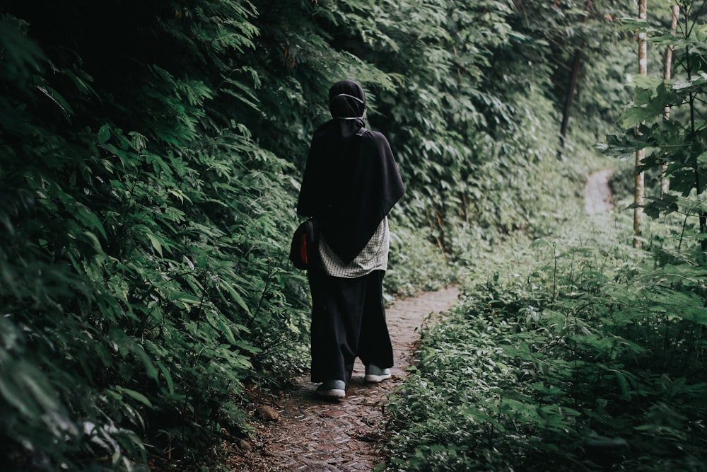 man in black jacket and black pants standing on pathway between green plants during daytime