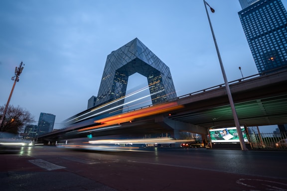 A modern architectural building is prominently featured with an angular, geometric design. The structure is surrounded by tall, urban skyscrapers, with a bridge in the foreground. Vehicles create streaks of light, indicating motion and a long exposure photograph.