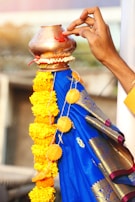A hand is applying a red mark on a copper vessel adorned with a garland of yellow marigold flowers. The vessel is placed on top of a blue fabric with golden patterns, likely a saree or drape. The background is blurred, focusing attention on the hand and the decorated vessel.
