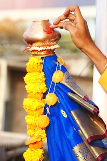 A hand is applying a red mark on a copper vessel adorned with a garland of yellow marigold flowers. The vessel is placed on top of a blue fabric with golden patterns, likely a saree or drape. The background is blurred, focusing attention on the hand and the decorated vessel.