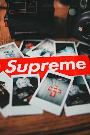 A collection of instant photographs scattered on a table, alongside two cameras. In the foreground, a prominent red Supreme sticker is visible. The images capture various scenes, some featuring clothing with the same logo.