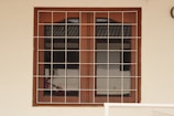 A wooden framed window with a metal grid installed over it. The window panes are reflective, displaying a faint reflection of a building structure and some foliage. The frame is set in a pale-colored wall, and there is a portion of a cream railing visible at the bottom.