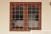 A wooden framed window with a metal grid installed over it. The window panes are reflective, displaying a faint reflection of a building structure and some foliage. The frame is set in a pale-colored wall, and there is a portion of a cream railing visible at the bottom.