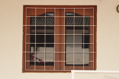 A wooden framed window with a metal grid installed over it. The window panes are reflective, displaying a faint reflection of a building structure and some foliage. The frame is set in a pale-colored wall, and there is a portion of a cream railing visible at the bottom.