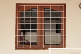 A wooden framed window with a metal grid installed over it. The window panes are reflective, displaying a faint reflection of a building structure and some foliage. The frame is set in a pale-colored wall, and there is a portion of a cream railing visible at the bottom.