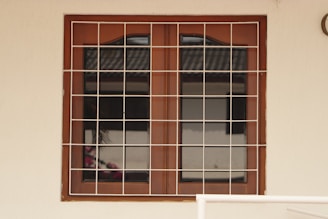A wooden framed window with a metal grid installed over it. The window panes are reflective, displaying a faint reflection of a building structure and some foliage. The frame is set in a pale-colored wall, and there is a portion of a cream railing visible at the bottom.