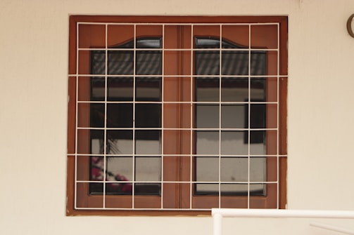 A wooden framed window with a metal grid installed over it. The window panes are reflective, displaying a faint reflection of a building structure and some foliage. The frame is set in a pale-colored wall, and there is a portion of a cream railing visible at the bottom.