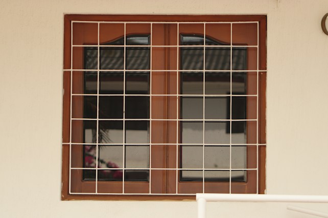 A wooden framed window with a metal grid installed over it. The window panes are reflective, displaying a faint reflection of a building structure and some foliage. The frame is set in a pale-colored wall, and there is a portion of a cream railing visible at the bottom.