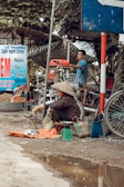 A street market scene with a woman wearing a traditional conical hat sitting on the ground, selling fish on an orange cloth. Next to her are two woven baskets and a green scale. A man, standing in the background, seems to be working with equipment on a bicycle cart. The environment appears rustic and busy with several signs and motorcycles around.