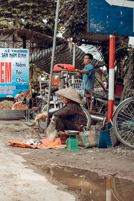 A street market scene with a woman wearing a traditional conical hat sitting on the ground, selling fish on an orange cloth. Next to her are two woven baskets and a green scale. A man, standing in the background, seems to be working with equipment on a bicycle cart. The environment appears rustic and busy with several signs and motorcycles around.