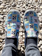 A pair of slip-on shoes featuring a colorful print with various images resembling cards or tiles, each with different designs and some text. The shoes are worn by someone wearing black socks and blue jeans, standing on a bed of light-colored, irregularly shaped stones.