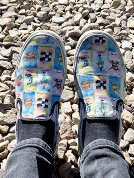 A pair of slip-on shoes featuring a colorful print with various images resembling cards or tiles, each with different designs and some text. The shoes are worn by someone wearing black socks and blue jeans, standing on a bed of light-colored, irregularly shaped stones.