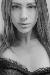 A grayscale portrait of a woman with long hair. Her expression is neutral and her gaze is fixed on the camera. She is wearing a lace-trimmed top.