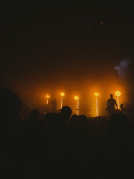 A dark concert scene illuminated by intense amber lights. Silhouettes of performers and audience members are visible in the dim lighting. Smoke or mist is also present, contributing to the moody atmosphere.