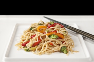 A plate of stir-fried noodles garnished with colorful vegetables such as broccoli, red and yellow bell peppers. The dish is presented on a white square plate with a pair of chopsticks resting beside it.